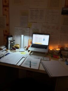 A focused language consultant workspace featuring a laptop, highlighted study frameworks, and linguistic deconstruction notes on the wall.